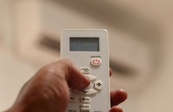 Asian man's hand sets the air conditioner temperature with a remote control in a bedroom before going to sleep during summer. Home cooling and nighttime comfort.