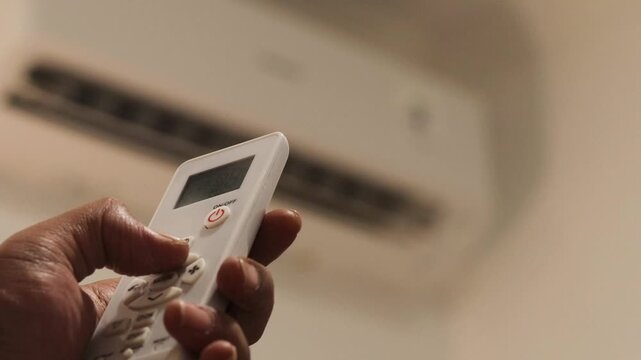 Asian man's hand sets the air conditioner temperature with a remote control in a bedroom before going to sleep during summer. Home cooling and nighttime comfort.