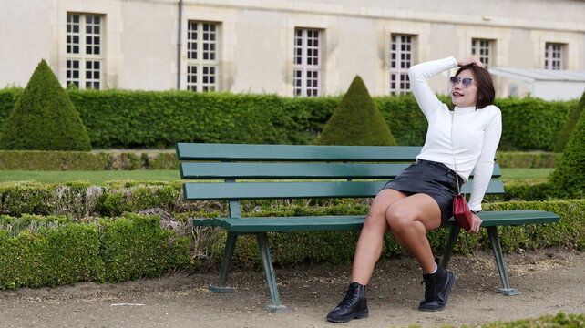 Stylish woman with short brown hair wearing a white long-sleeve ribbed top, black leather skirt and black  boots, poses confidently on a green bench in a park