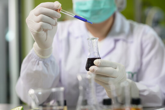 Scientist in a lab coat uses a dropper to transfer blue liquid into a flask with dark solution, performing an experiment in a laboratory setting.