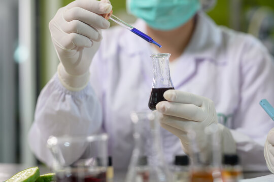Scientist wearing gloves and a mask uses a dropper to transfer a blue liquid into a flask containing a dark liquid in a laboratory setting.