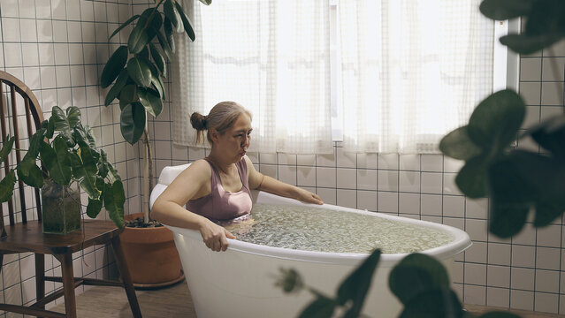 A mature Asian woman takes an ice bath with arms crossed in a bright, plant-filled bathroom with white tiles, focusing on cold therapy recovery.
