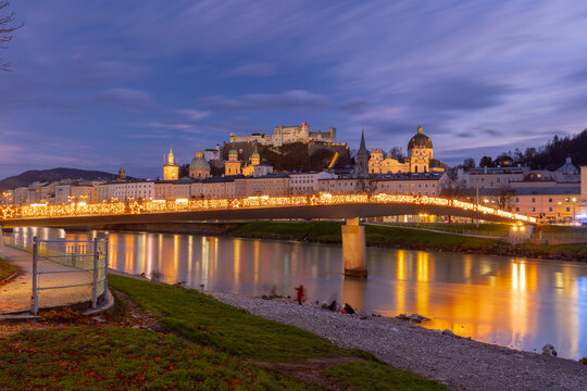 Salzburg Old Town with Hohensalzburg Fortress and an illuminated bridge reflected in the Salzach River at blue hour, Austria.