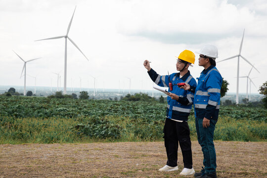 Two engineers in hard hats and safety gear reviewing documents on a clipboard at a wind turbine farm.