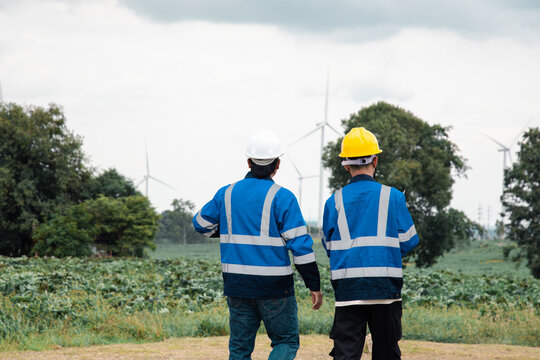 Two engineers in hard hats and safety gear reviewing documents on a clipboard at a wind turbine farm.