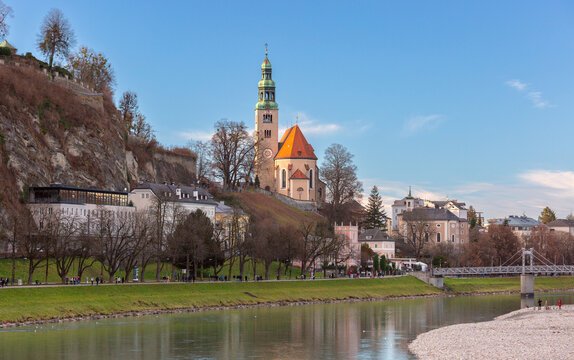 View of Mullner Church above the Salzach River with historic buildings and riverside promenade in Salzburg, Austria.