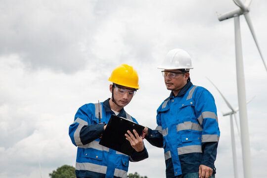Two engineers in hard hats and safety gear reviewing documents on a clipboard at a wind turbine farm.