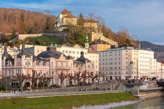 Historic buildings and riverside promenade at the foot of Monchsberg hill in Salzburg, Austria.