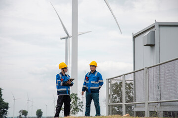 Fototapeta premium Two engineers in hard hats and safety gear reviewing documents on a clipboard at a wind turbine farm.