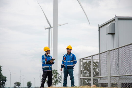 Two engineers in hard hats and safety gear reviewing documents on a clipboard at a wind turbine farm.