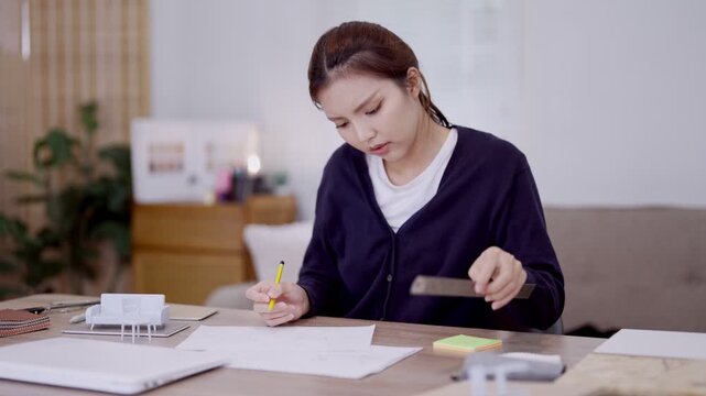 Young Asian furniture designer writing notes and sketching design plans on paper at modern home studio