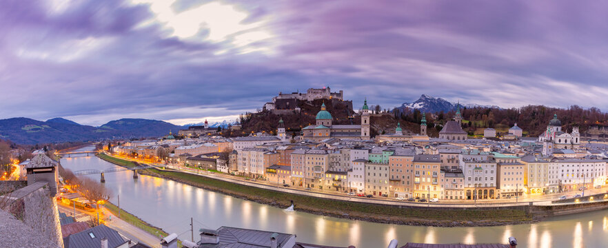Wide panoramic view of Salzburg Old Town along the Salzach River with historic architecture and Hohensalzburg Fortress at dusk, Austria.