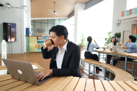 Asian Businessman Drinking Coffee While Working on Laptop in Modern Office