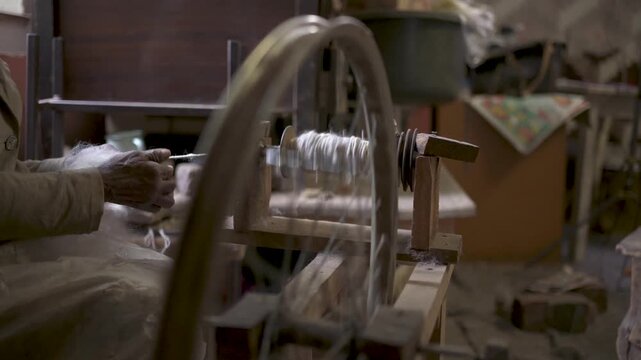 Elderly woman spinning wool into yarn using traditional wooden spinning wheel.