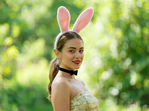 A young woman wearing a pink bunny ear headband poses outdoors in a scenic garden