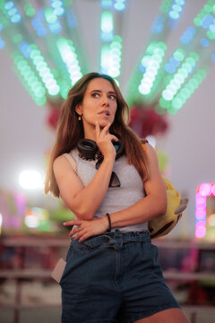 Thoughtful Woman With Headphones Standing in the Amusement Park. Pensive attractive girl with headphones around neck standing in front of colorful bokeh lights 