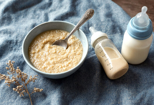 Homemade baby cereal in a bowl with a spoon and baby bottles on a blue cloth background