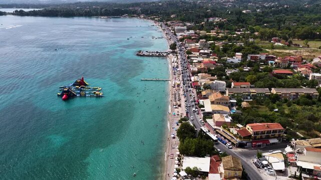 Aerial flight over busy Ipsos Beach in Corfu Greece showing turquoise water, coastal road, resorts and a fun water park