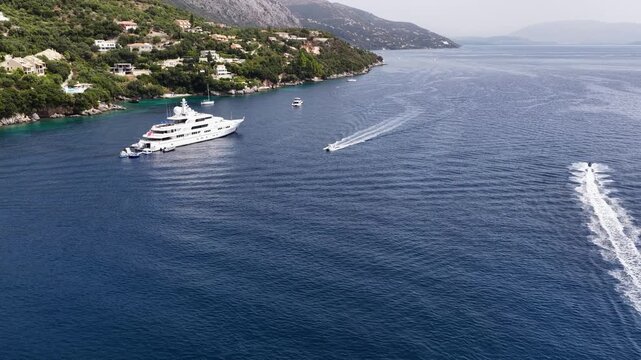 Aerial drone view of luxury superyacht anchored in blue sea with speedboat creating wake near Barbati, Corfu island, Greece.