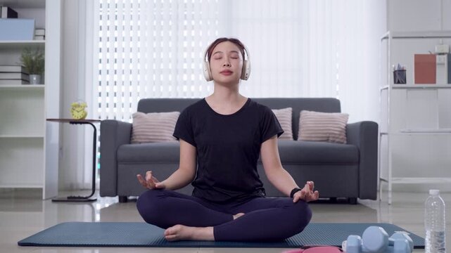 Young Asian woman meditating in lotus pose wearing headphones on yoga mat in bright modern living room