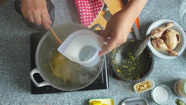 Overhead view of pouring water from measuring pitcher into pot with saut&eacute;ed spices on the stove
