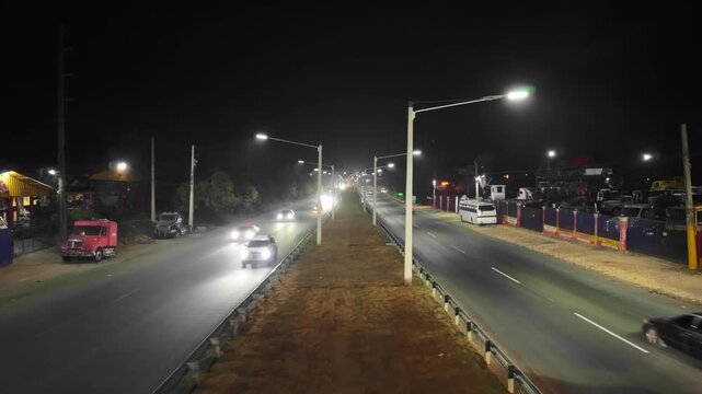 Static high-angle aerial drone shot overlooking a multi-lane highway at night, capturing dump trucks, buses, cars, and motorcycles driving under street lights
