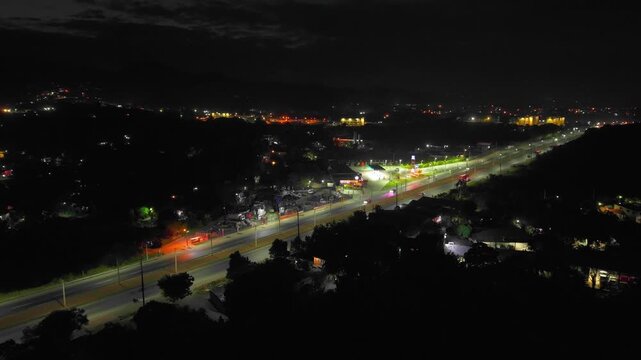 High-angle aerial drone shot ascending and flying backward over an illuminated highway at night, capturing continuous vehicle traffic driving past lit gas stations