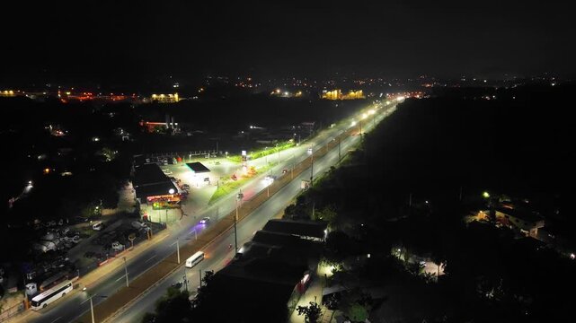 High-angle aerial drone shot ascending and panning right over an illuminated highway at night, capturing vehicle traffic driving past a glowing gas station and local businesses