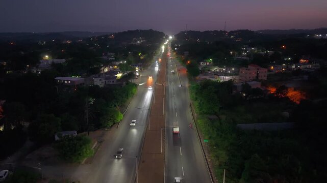 High-angle aerial drone shot advancing over an illuminated highway at dusk, capturing steady vehicle traffic driving past a gas station, local businesses, and distant hills