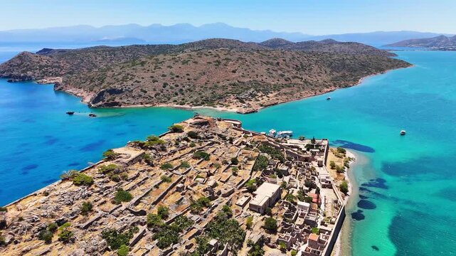 Kalydon ruins recede beneath an aerial view off Crete, exposing stone foundations, a perimeter wall, narrow beach and turquoise channels framed by arid islands and distant mountains.