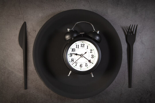 Alarm clock on black plate with fork and knife, top view