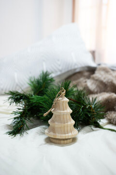 A natural wooden Christmas tree ornament sits gracefully on white bedding next to a fresh pine branch. The soft morning light and cozy textures create a serene, minimalist holiday atmosphere.