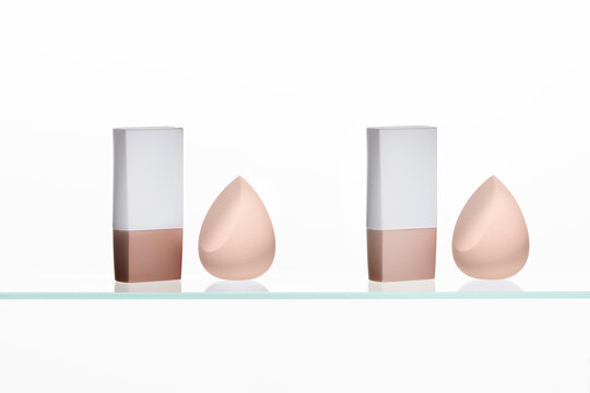 Two colors foundation stick and a perfectly matched nude makeup sponge on a clear glass shelf against a bright, clean white background. Essential base tools.