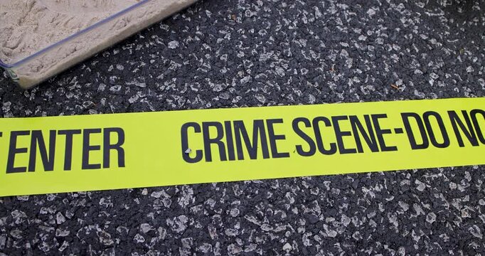 Slow motion footage showing bright yellow crime scene do not enter barrier tape stretched across a rough tarmac road surface creating a dramatic close-up view of a restricted area under investigation