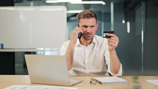 Professional man makes phone call for online payment support. Holds credit card with worried expression, financial transaction difficulty. Trouble with digital banking process or credit card security.