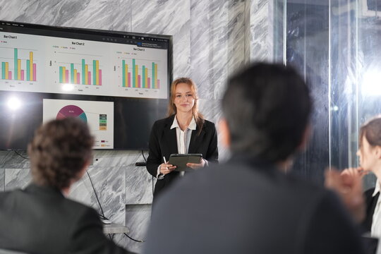 Blonde businesswoman gestures with a tablet in hand while presenting colourful charts on a wall screen to a seated audience in a modern marble boardroom of a Bangkok office.
