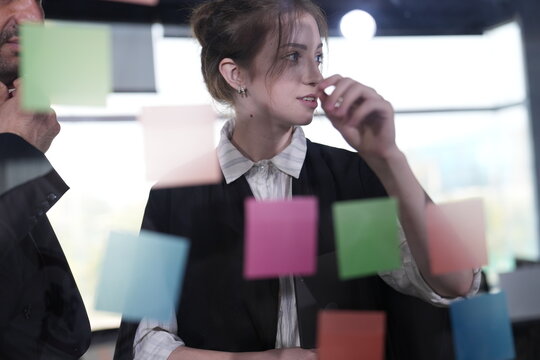 Close view of a pensive young woman holding her hand near her mouth while she rereads the sticky notes arranged across the glass wall of a modern coworking office.