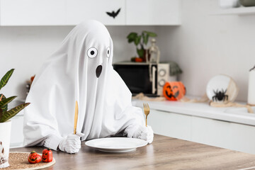 Child in ghost costume holding a fork and knife at a dining table with halloween decorations in a...