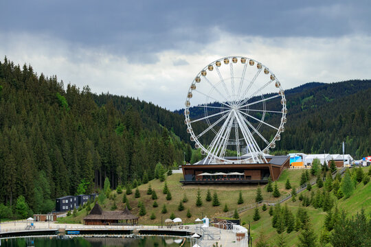 A large observation wheel stands prominently on a grassy hill surrounded by dense evergreen forests and rolling hills