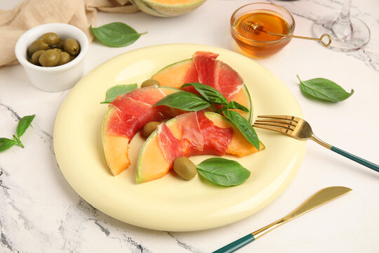 Plate of fresh melon pieces with delicious prosciutto, olives and basil leaves on white background, closeup