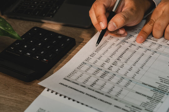 Accountant checking financial report figures with pen beside calculator and laptop for accounting calculation, audit, tax preparation, bookkeeping.