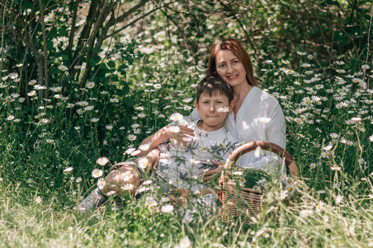 Caucasian mother and son resting together within a sunlit field filled with vibrant daisies during a warm summer afternoon