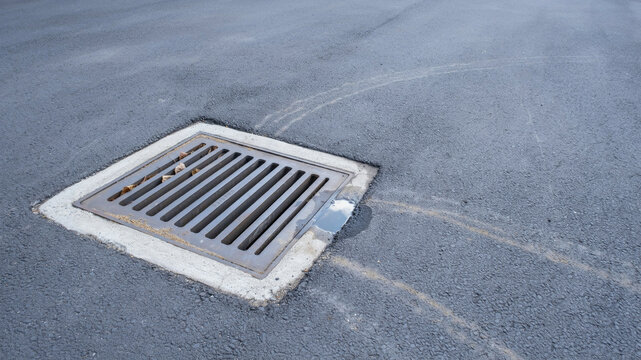 Square metal grate drain cover set into asphalt pavement with visible tire marks and a small puddle of water on the right side