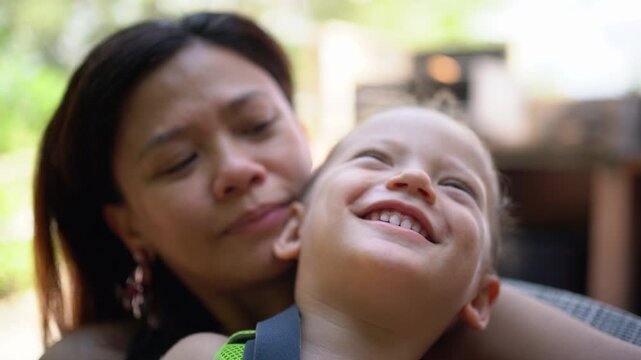 A mother taps her fingers on her son's mouth to make a playful sound as they both laugh together outdoors