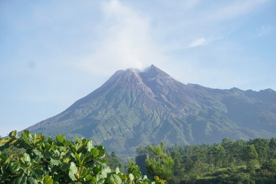 Scenic landscape photo of Mount Merapi, an active volcano in Central Java, Indonesia. The tall conical mountain peak releases smoke and steam into a clear blue sky. 