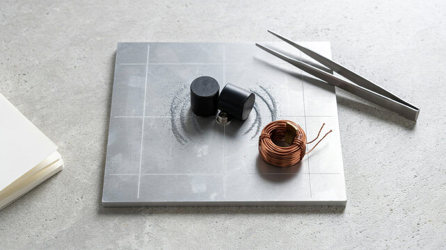 Two cylindrical black magnets and a coil of copper wire on a metallic surface with tweezers and a stack of paper, suggesting a science experiment or electrical engineering concept.keywords