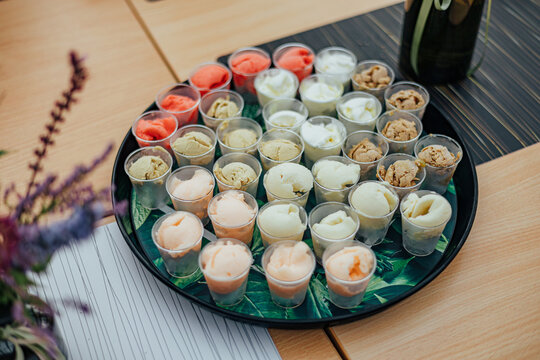 Tray filled with small cups of assorted ice cream samples in multiple flavors and colors, presented on a table with decorative flowers for a tasting event