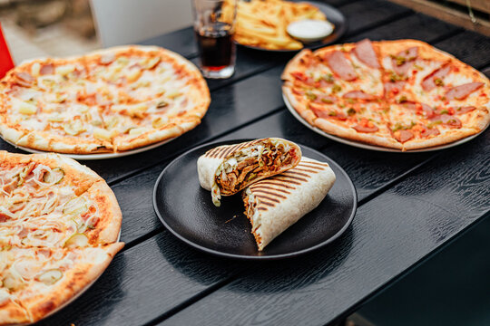 Grilled wrap cut in half and filled with meat and cabbage, served on a black plate among several pizzas, fries and a soft drink on an outdoor dining table