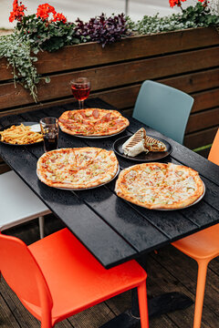 Colorful outdoor restaurant table arranged with several pizzas, a grilled wrap, French fries and drinks, surrounded by modern chairs and wooden planters