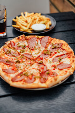 Freshly baked salami pizza topped with cheese and diced peppers on a dark restaurant table, with French fries, dipping sauce and a soft drink in the background